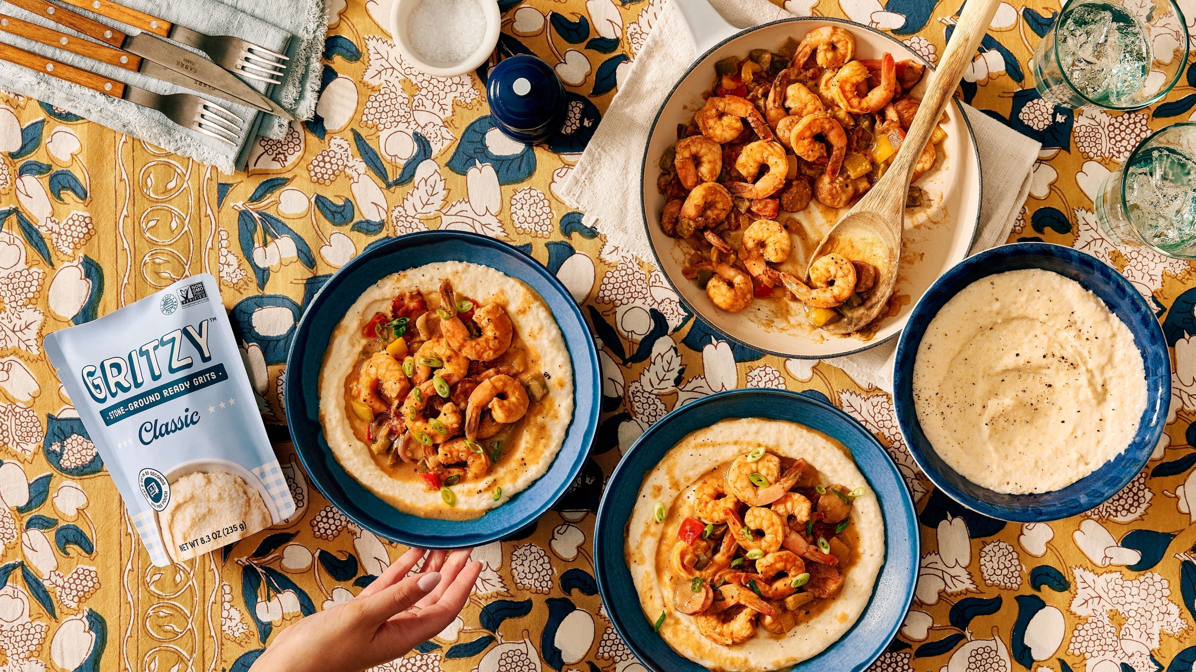 Overhead table spread of Southern shrimp and grits in blue bowls with a skillet of Cajun shrimp, andouille sausage, and peppers alongside a Gritzy Classic grits pouch